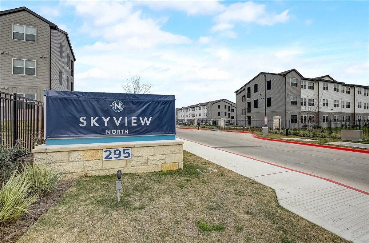 Landscaped community entrance with stone monument feature, sidewalk, and internal roadway leading to apartment homes.